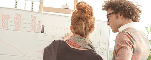 Junge Frau und junger Mann stehen vor einem Whiteboard und betrachten darauf gezeichnete Diagramme. Junge Frau und junger Mann stehen vor einem Whiteboard und betrachten darauf gezeichnete Diagramme.