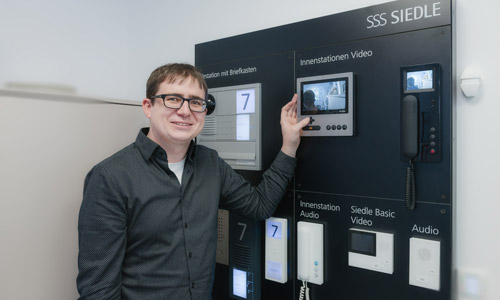 Philipp Denz, ein Mann mit Brille, vor einer Wand mit verschiedenen Modellen von Sprechanlagen. Philipp Denz, ein Mann mit Brille, vor einer Wand mit verschiedenen Modellen von Sprechanlagen.
