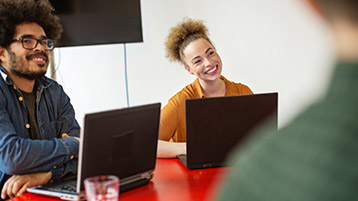 Ein junger Mann und eine junge Frau sitzen an Laptops an einem roten Tisch.