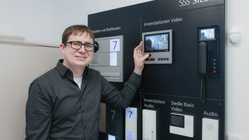 Ein Mann mit Brille steht vor einer Wand mit verschiedenen Modellen von Sprechanlagen.