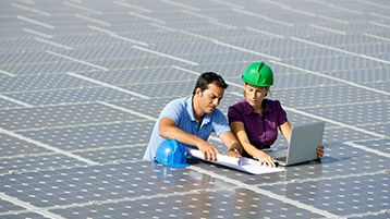 Mann mit Bauplänen und Frau mit Bauhelm auf Solarfeld, Frau bedient Laptop