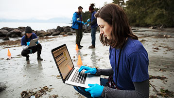 Junge Menschen in Arbeitskleidung sind mit Laptops an einem nassen Strand.