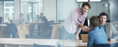 Ein junger Mann im rosafarbenen Hemd, aufstehend, schüttelt einem anderen jungen Mann, sitzend, über einen Schreibtisch hinweg die Hand. Mit am Tisch sitzt ein dritter Mann, lächelt. Ein junger Mann im rosafarbenen Hemd, aufstehend, schüttelt einem anderen jungen Mann, sitzend, über einen Schreibtisch hinweg die Hand. Mit am Tisch sitzt ein dritter Mann, lächelt.
