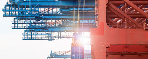 Im Vordergrund ein roter, im Hintergrund mehrere blaue Containerkräne. An einem davon hängt ein Container im Gegenlicht. Im Vordergrund ein roter, im Hintergrund mehrere blaue Containerkräne. An einem davon hängt ein Container im Gegenlicht.