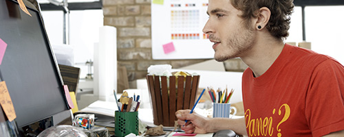 Ein junger Mann im roten T-Shirt arbeitet am Schreibtisch am Computer. Ein junger Mann im roten T-Shirt arbeitet am Schreibtisch am Computer.